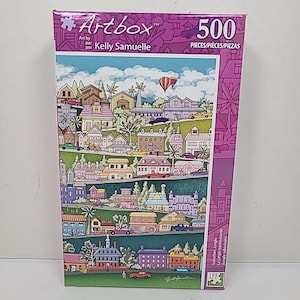 Peut inclure: Puzzle de 500 pièces dans une boîte violette. L'illustration représente une banlieue colorée avec des maisons, des voitures et une montgolfière. La boîte porte le logo "Artbox" et le nom de l'artiste, Kelly Samuelle.