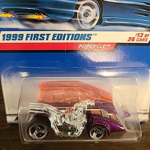 Puede incluir: Un coche de juguete Popcycle Hot Wheels de la serie First Editions de 1999. El coche presenta un motor cromado, carrocería morada y una capota naranja. El embalaje indica "#13 of 26 Cars".