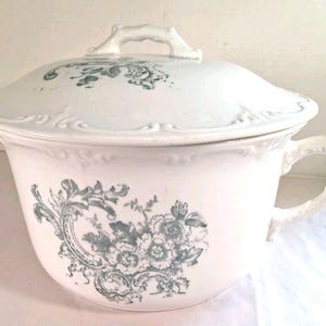 Op de afbeelding: Witte keramische po met deksel en handvat. De pot heeft een bloemenontwerp in blauwgroene tinten. Het deksel heeft een handvat en decoratieve rand. Een klassiek stuk voor de interieurdecoratie.
