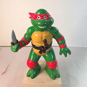 May include: A green Teenage Mutant Ninja Turtle figurine with a red eye mask, red elbow and knee pads, and a yellow chest plate. The character wears a black belt with a red letter R and holds a silver-colored sai in its right hand.