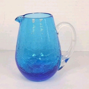 May include: A blue glass pitcher with a crackle finish. The pitcher has a clear handle and a spout for pouring. The color transitions from light blue at the top to a deep blue at the base. A decorative piece for serving beverages.