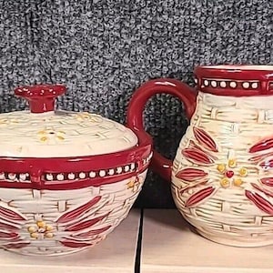 以下が含まれることがあります： 赤と黄色の花柄が施された白いセラミック製の砂糖入れとクリーマーのセット。セットには、編まれたバスケット模様と赤い縁取りが施されています。