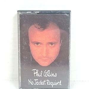 以下が含まれることがあります： フィル・コリンズのアルバム「No Jacket Required」をフィーチャーしたヴィンテージカセットテープ。アルバムのカバーには、フィル・コリンズの顔のクローズアップが表示されています。カセットは透明なプラスチックケースに入っています。