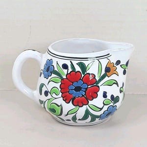 May include: White ceramic creamer with a handle and spout, adorned with a vibrant floral design. The flowers are red, blue, and orange, with green leaves and black accents. A decorative piece for serving cream or other liquids.