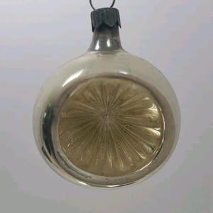 May include: A vintage Christmas ornament with a silver-colored exterior and a gold-colored, starburst-patterned interior. The ornament has a metal cap and a loop for hanging. The overall shape is round.
