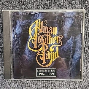 May include: CD case featuring The Allman Brothers Band logo in gold script over a dark denim background. The text "a decade of hits 1969-1979" is in a white rectangle at the bottom. The CD case is in a clear plastic case.