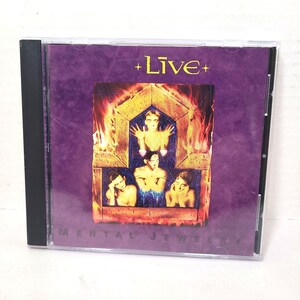 Op de afbeelding: Een cd-hoesje met het album "Mental Jewelry" van de band Live. De hoes toont figuren in een vurige, goudkleurige structuur tegen een dieppaarse achtergrond. De bandnaam "+Live+" is in het wit gedrukt. De albumtitel staat ook in het wit.