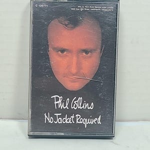 以下が含まれることがあります： フィル・コリンズのアルバム「No Jacket Required」のヴィンテージカセットテープ。アルバムのカバーには、白い文字でアルバムタイトルが書かれたフィル・コリンズの肖像画が描かれています。カセットケースは透明なプラスチック製です。
