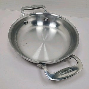 Pode incluir: Uma frigideira All-Clad em aço inoxidável com duas alças curvas. A panela tem uma superfície de cozimento circular e é feita de um metal brilhante e reflexivo. As alças são rebitadas à panela e apresentam o logotipo All-Clad.