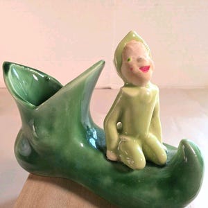Könnte beinhalten: Ein glänzender grüner Keramikübertopf in Form eines spitzen Schuhs mit einer kleinen, hellgrünen Elfenfigur, die auf der Schuhspitze kniet. Der Elf hat ein bemaltes Gesicht mit rosa Wangen und roten Lippen sowie eine passende spitze Mütze.
