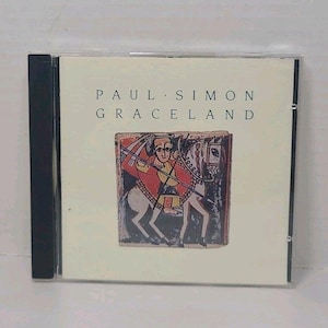 Könnte beinhalten: Eine CD-Hülle mit dem Album "Graceland" von Paul Simon. Das Cover zeigt eine farbenfrohe Illustration einer Person auf einem Pferd. Die CD-Hülle hat einen schwarzen Rücken und eine transparente Kunststofffront.