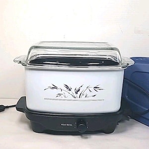 May include: White slow cooker with a clear glass lid and black base. The white body features a black floral design. A blue lid is visible on the right. The control knob reads "West Bend".