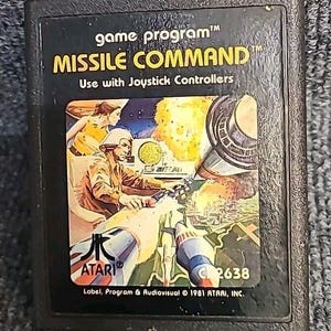 May include: A vintage Atari game cartridge for Missile Command. The cartridge is black with a colorful image of a spaceship and a city under attack. The text "game program™ MISSILE COMMAND™ Use with Joystick Controllers" is printed on the front of the cartridge. The Atari logo and the number C2638 are also printed on the front.