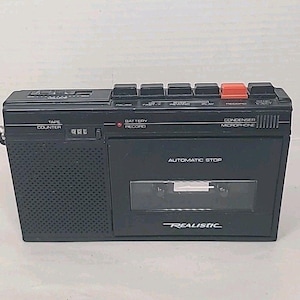 以下が含まれることがあります： 黒いRealisticカセットテープレコーダー。スピーカー、テープカウンター、各種ファンクションボタンを内蔵。オレンジ色の録音ボタンがあり、前面には「Automatic Stop」の文字が印刷されています。