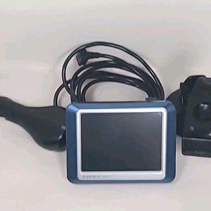 Puede incluir: Un dispositivo GPS Garmin con marco azul y plateado, pantalla negra y cable de alimentación negro. Incluye un cargador de coche y un soporte de montaje. El texto "GARMIN 1300" es visible.