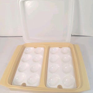 Könnte beinhalten: Ein gelber Eierbehälter aus Kunststoff mit einem durchsichtigen Deckel. Der Behälter hat zwei weiße Tabletts, jedes mit sechs eiförmigen Vertiefungen. Der Deckel ist geöffnet und gibt den Innenraum frei. Der Behälter ist für ein Dutzend Eier konzipiert.
