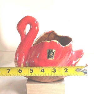 May include: A red ceramic swan-shaped planter with a glossy finish and a gold-toned sticker featuring a crown logo and the text A CAMERON QUALITY GIFT HAND MADE. A yellow measuring tape shows the object is approximately 19 cm in width.
