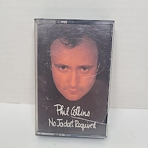 以下が含まれることがあります： フィル・コリンズのアルバム「No Jacket Required」のヴィンテージカセットテープ。アルバムのカバーには、フィル・コリンズの顔のクローズアップが写っています。カセットテープは透明なプラスチックケースに入っています。