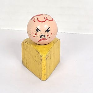 Pode incluir: Um brinquedo de madeira vintage com uma cabeça redonda rosa, cabelo encaracolado vermelho e uma expressão zangada. A cabeça assenta sobre um bloco triangular amarelo. O brinquedo mostra sinais de desgaste.
