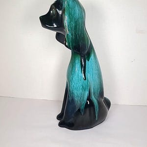 May include: A ceramic dog figurine with a glossy finish. The dog is primarily black with teal and green dripping glaze on the head and body. The dog is sitting with its head turned to the side.