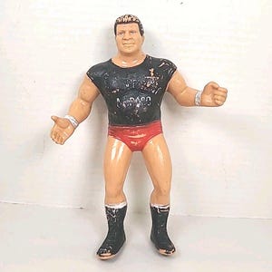 May include: A vintage action figure of a muscular man in a black t-shirt, red briefs, and black boots. The figure has tan skin, short brown hair, and white wristbands. The t-shirt has faded white text.