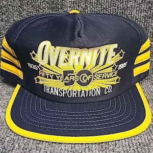 Może przedstawiać: Granatowo-żółta czapka typu trucker z żółtym napisem "OVERNITE" oraz "FIFTY YEARS OF SERVICE TRANSPORTATION CO.". Czapka ma żółty daszek i żółte paski po bokach. Widoczne są również lata 1935 i 1985.