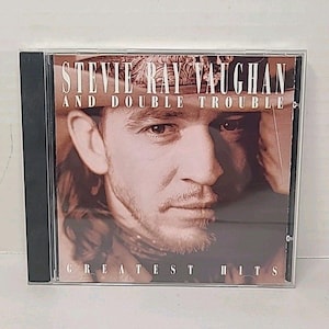 Pode incluir: Uma caixa de CD com o álbum Greatest Hits de Stevie Ray Vaughan and Double Trouble. A capa apresenta um retrato em sépia de Stevie Ray Vaughan. O texto na capa diz "Stevie Ray Vaughan and Double Trouble" e "Greatest Hits."