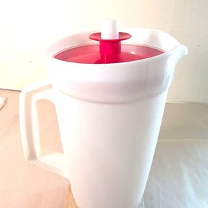 Può includere: Una caraffa in plastica bianca dotata di manico laterale e beccuccio. Il coperchio è rosso con un meccanismo a stantuffo cilindrico bianco centrato sulla parte superiore. La caraffa presenta una finitura liscia e opaca e una forma cilindrica leggermente conica.