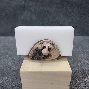 May include: A whimsical, handcrafted ceramic sculpture of a bearded face with a large nose and detailed facial features. The sculpture is a light brown color and is mounted on a wooden base. The sculpture is partially obscured by a white block.