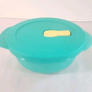 May include: A teal-colored, round food storage container with a matching lid. The lid features a yellow tab for easy opening and closing. The container has two side handles for carrying.