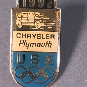 May include: Vintage 1992 Chrysler Plymouth USA Olympic pin. The pin features a gold-colored frame with a black top section displaying the year. Below, a car graphic is above the text "CHRYSLER Plymouth". The bottom section is blue with "USA" and Olympic rings.