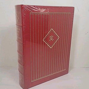 Puede incluir: Un libro de tapa dura rojo, envuelto en plástico. La portada presenta líneas verticales doradas y un emblema en forma de diamante. El lomo tiene texto y diseños rectangulares dorados. El libro está de pie.