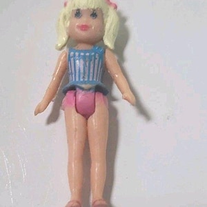 May include: A small plastic doll with blonde hair styled in pigtails, adorned with pink bows. The doll wears a blue and white striped top, pink shorts, and pink shoes. The doll's arms are outstretched.