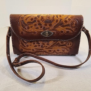 Puede incluir: Bolso bandolera de cuero marrón con intrincados grabados florales y de volutas. El bolso tiene un cierre de solapa con un broche de metal y una correa larga y ajustable. El cuero tiene un efecto de dos tonos, con áreas más claras.