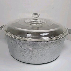 Puede incluir: Una olla de cocina de color plateado con una tapa de vidrio transparente y un pequeño pomo. La olla tiene dos asas laterales y una superficie texturizada. La tapa es transparente.