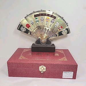 May include: A decorative fan with a gold-colored frame and a black base, featuring colorful painted faces. The fan is displayed on a red box with gold accents and a clasp. The fan is approximately 12 inches wide.