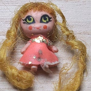 May include: A small vintage doll with yellow yarn hair styled in pigtails. The doll has a pink dress with a white trim and a decorative bodice. The doll's face has painted features, including large green eyes and rosy cheeks.