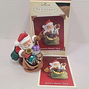 Könnte beinhalten: Ein Hallmark Keepsake Ornament mit dem Weihnachtsmann und einem Sack voller Spielzeug. Die Figur ist rot, weiß und braun, mit dem Text "Santa's Magic Sack" auf der Verpackung und der Karte. Das Ornament ist ein VIP-Geschenk von 2005.