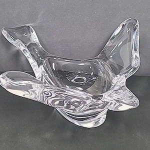 May include: A clear glass decorative bowl with a unique, abstract design. The bowl has a flowing, organic shape with curved edges and a central depression. The glass is transparent, allowing light to pass through, and sits on a dark surface.