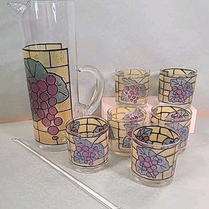 Puede incluir: Un juego de seis vasos transparentes de vidrio y una jarra con un diseño de vidriera de uvas y hojas. Los vasos y la jarra tienen un fondo amarillo, dorado y marrón con un contorno negro. Las uvas son moradas y las hojas son verdes.