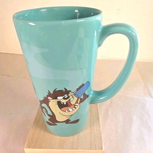 May include: A tall, light blue ceramic mug featuring a graphic of the Tasmanian Devil character holding a blue object. The mug has a tapered shape, a wide rim, and a curved handle on the right side, with a light gray swirl design behind the character.
