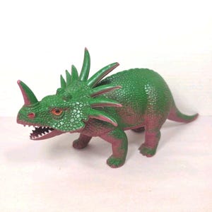 May include: A green and purple dinosaur figurine with a textured, scaly surface. The creature features a prominent horn on its snout, a frill with multiple spikes extending from the back of its head, and orange eyes with an open mouth showing teeth.
