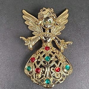 May include: Gold-toned angel brooch with outstretched arms and detailed wings. The angel's dress is adorned with red and green gemstones. The brooch has a vintage aesthetic, perfect for adding a touch of elegance to any outfit.