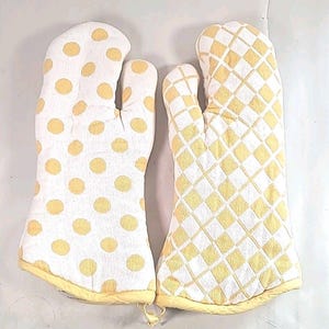 May include: A pair of oven mitts. One mitt features a white background with yellow polka dots. The other mitt has a white background with a yellow diamond pattern. Both mitts have yellow trim and a loop for hanging.