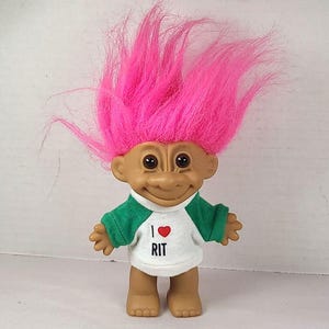 Puede incluir: Una muñeca troll vintage con cabello rosa brillante y una camisa blanca con mangas verdes. La camisa tiene el texto "I ❤️ RIT" con un corazón rojo. La muñeca tiene ojos marrones y está de pie.