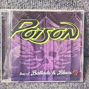 May include: A compact disc case with a purple and black background featuring the band Poison's logo in yellow and black. The text "Best of Ballads & Blues" is printed in white on a black banner.