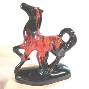 May include: A glossy ceramic horse figurine standing on a solid black base. The sculpture features a striking color combination of deep black and vibrant red, with the red glaze appearing in a streaked, flame-like pattern across the horse's body.