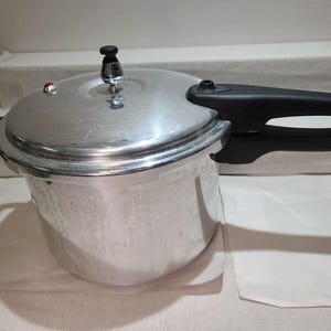 Op de afbeelding: Een zilveren snelkookpan met een zwarte handgreep en deksel. De pan heeft een glanzende metalen afwerking en een zwarte knop bovenop. De handgreep is gebogen en aan de zijkant van de pan bevestigd. De pan staat op een wit oppervlak.