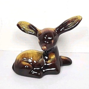 May include: A ceramic deer figurine in shades of brown and yellow. The deer is in a resting position with its ears upright. The figurine has a glossy finish and is likely a decorative item.