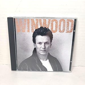Puede incluir: Una caja de CD con una imagen en blanco y negro de un hombre, con el nombre "WINWOOD" en letras naranjas. El hombre lleva una chaqueta negra sobre una camisa blanca. La caja del CD es transparente.